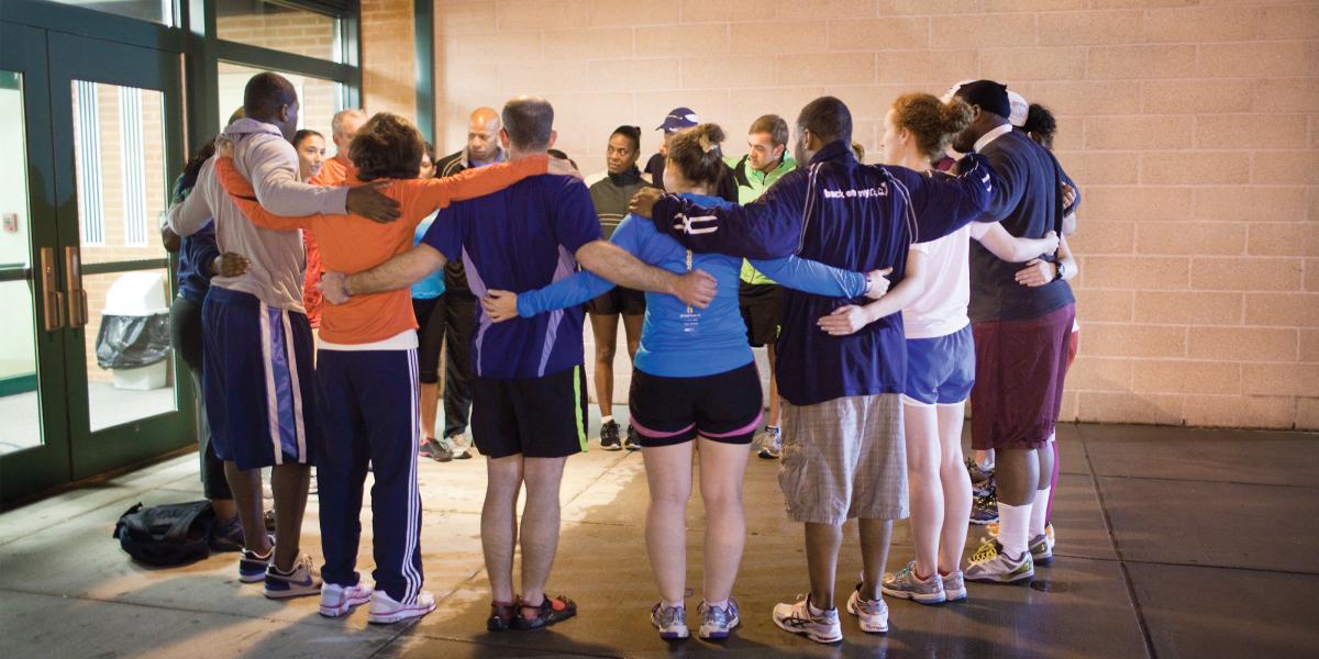 a circle of about 15 people in athletic gear stand in a circle with their arms around each other