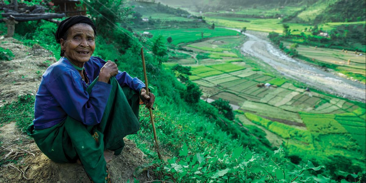 An older Nepali woman smiles and sits on a hill overlooking a field