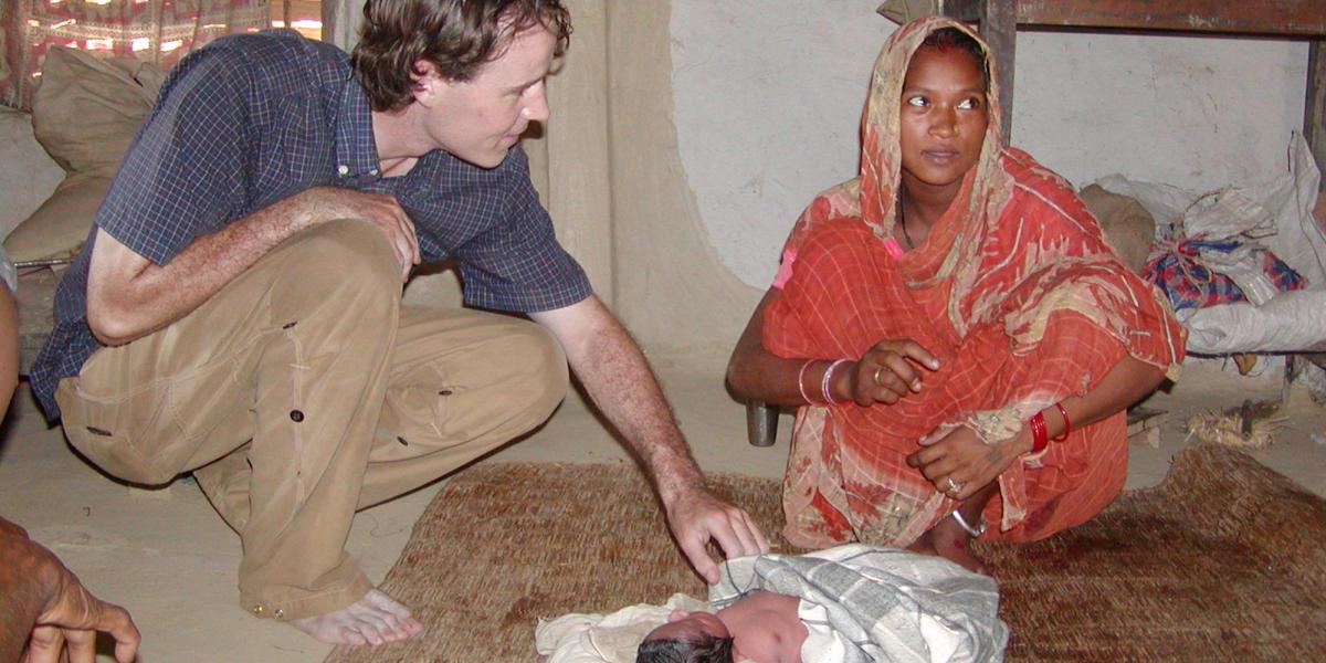 A researcher squats on the floor to examine a newborn infant; the child's mother, in a red sari, sits next to the infant.