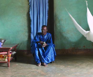 Ogobaro Doumbo sits on a low chair in his home. A mosquito net hangs to his left.